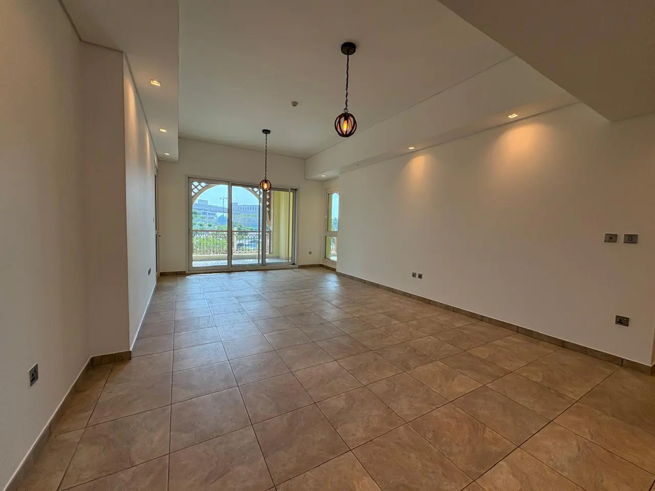 Empty room with beige tile floor, white walls, and sliding glass doors to a balcony. Two hanging light fixtures are visible.
