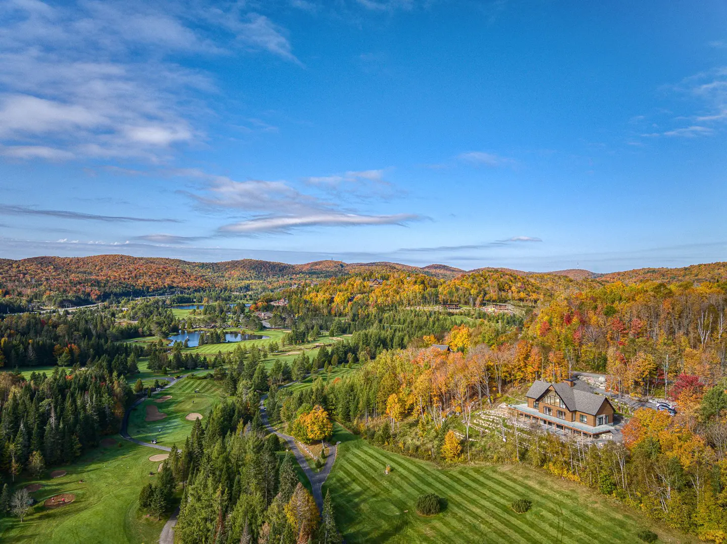 Aerial view of a brown house with a deck, surrounded by fall foliage and a golf course under a blue sky.