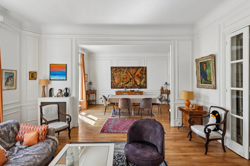 Bright, airy living room with hardwood floors, white walls, and ornate molding. A dining room is visible through an open doorway. Artwork adorns the walls.