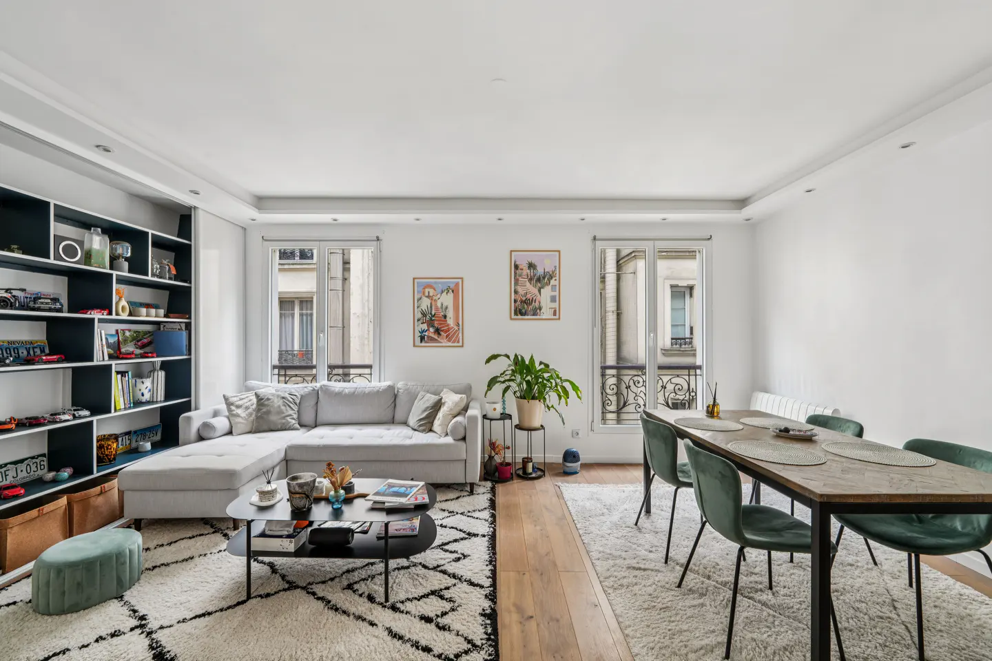 Bright living room with white walls, a gray sectional sofa, a black coffee table, and a dining table with green chairs.