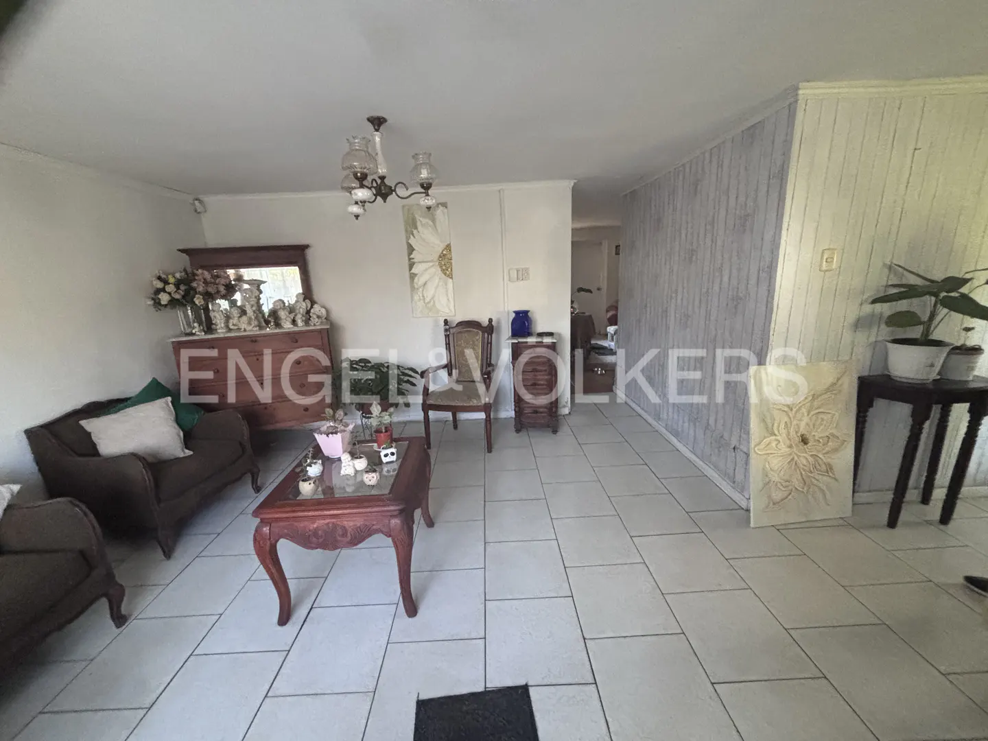 Living room with tile floors, brown sofas, and a wooden coffee table. A dresser with flowers sits against the wall. A chandelier hangs from the ceiling.