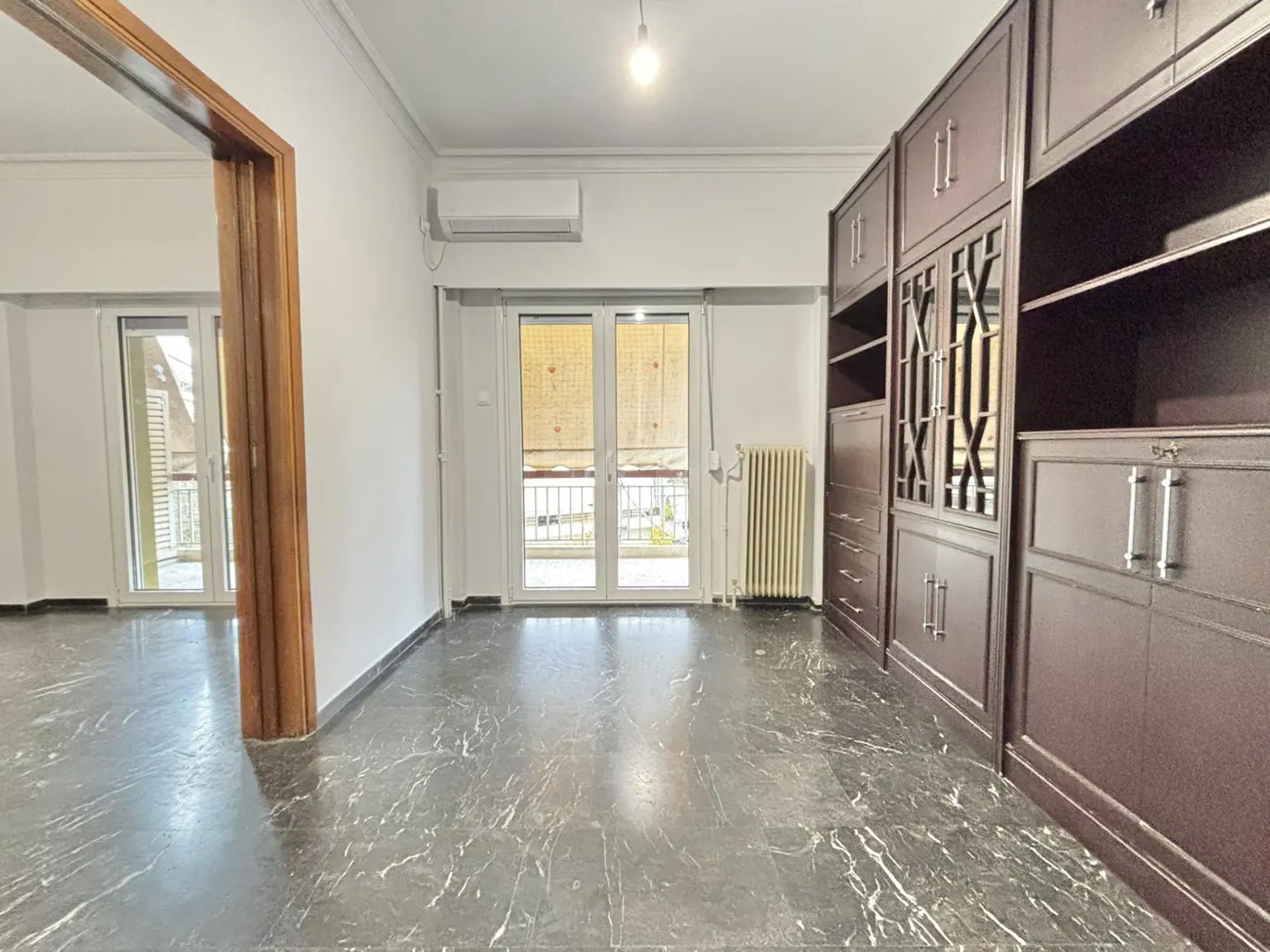 A bright room with a marble floor, white walls, and a large, dark wood built-in cabinet. A balcony door lets in natural light.