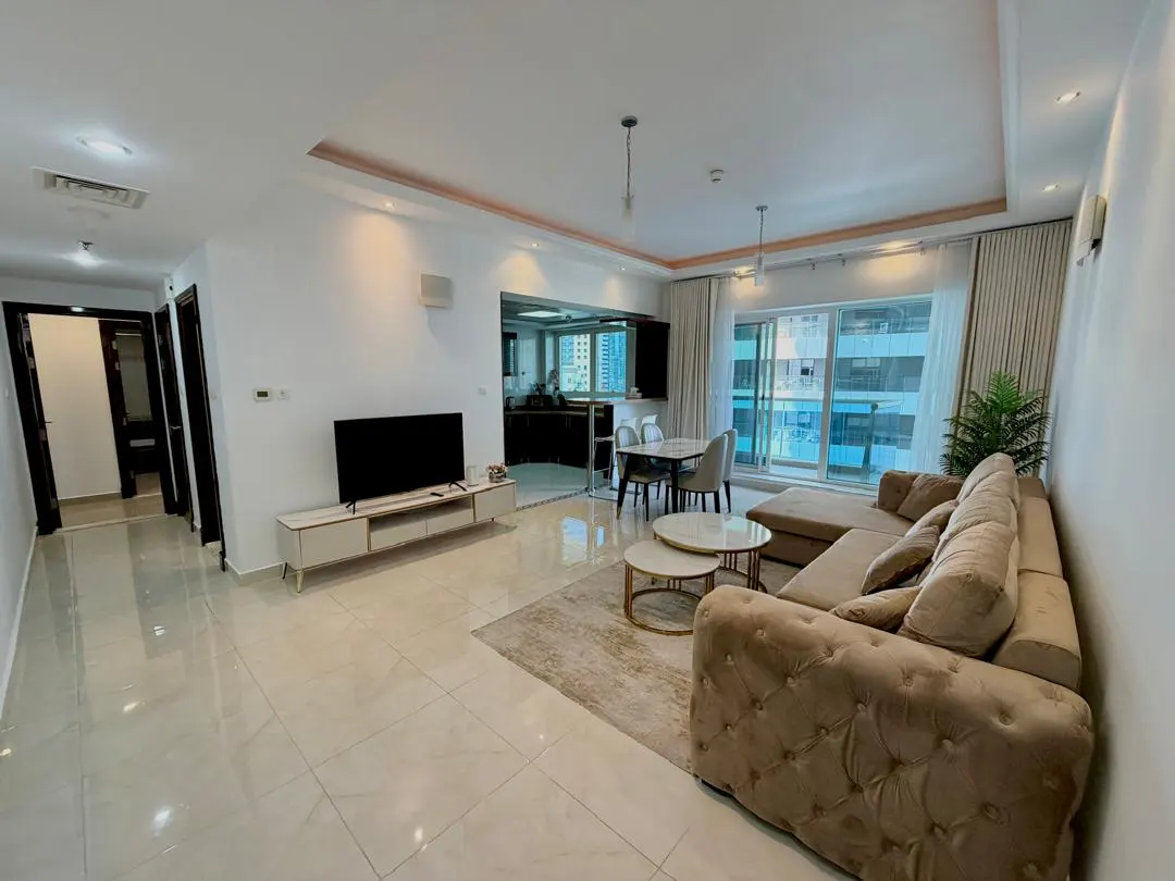 Modern open-plan living room with beige tufted sofa, coffee tables, TV console, dining area, and large floor-to-ceiling windows.