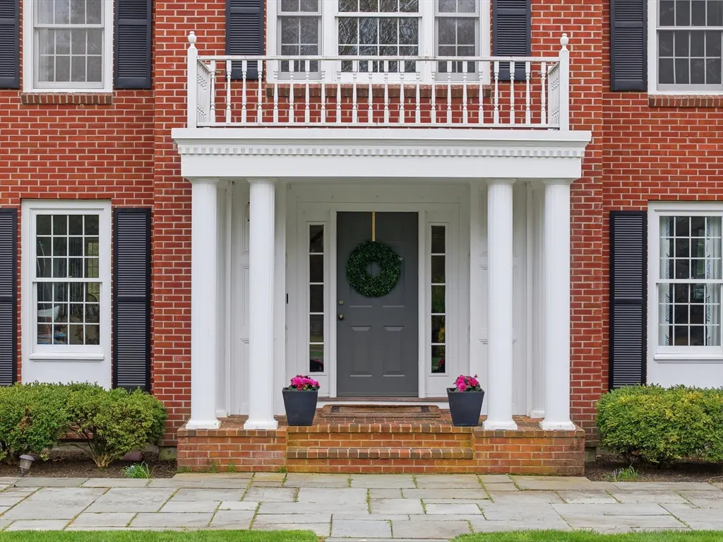 Red brick house with white columns framing a gray front door with a green wreath. Black shutters accent the windows.