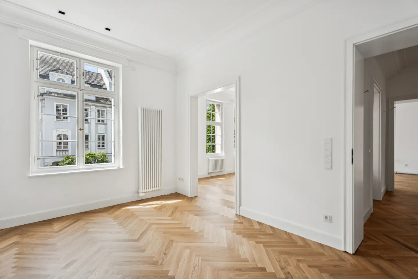 Bright, empty room with herringbone wood floors, white walls, and a large window showing a building exterior.
