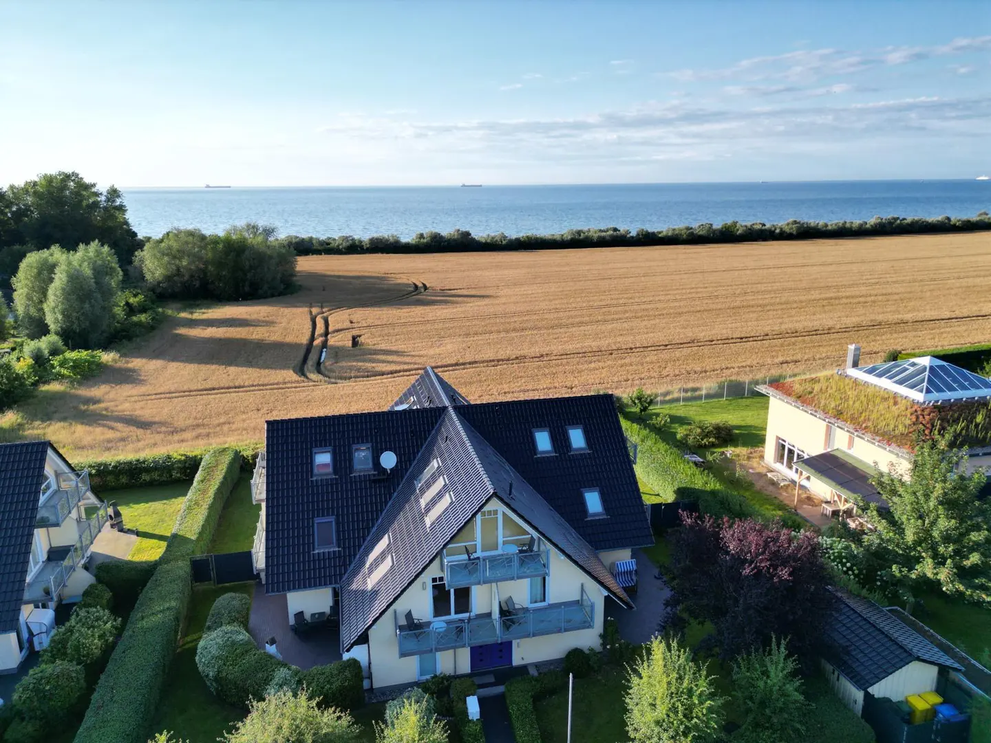 Aerial view of a yellow house with a black roof, balconies, and dormer windows, near a wheat field and the ocean.