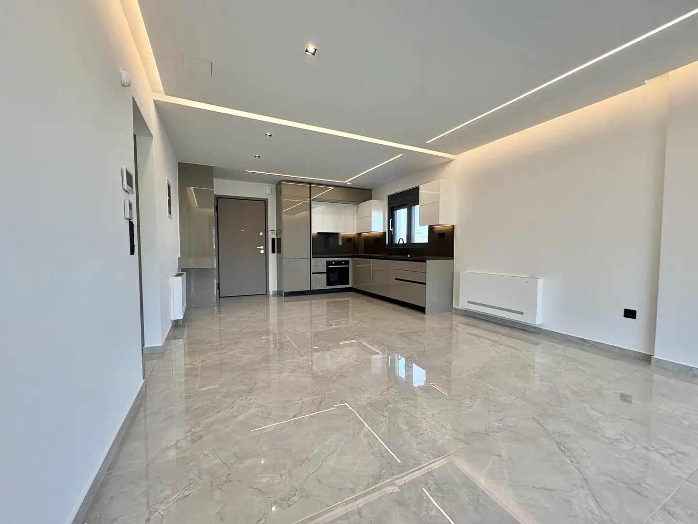 A modern apartment interior with gray marble floors, white walls, and a gray and white kitchen. Linear lighting is installed on the ceiling.