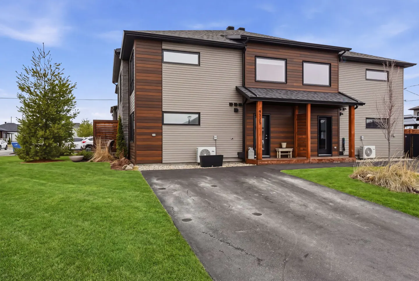 Modern two-story duplex with brown wood and gray siding, black trim, and a shared driveway. Green lawn and blue sky.