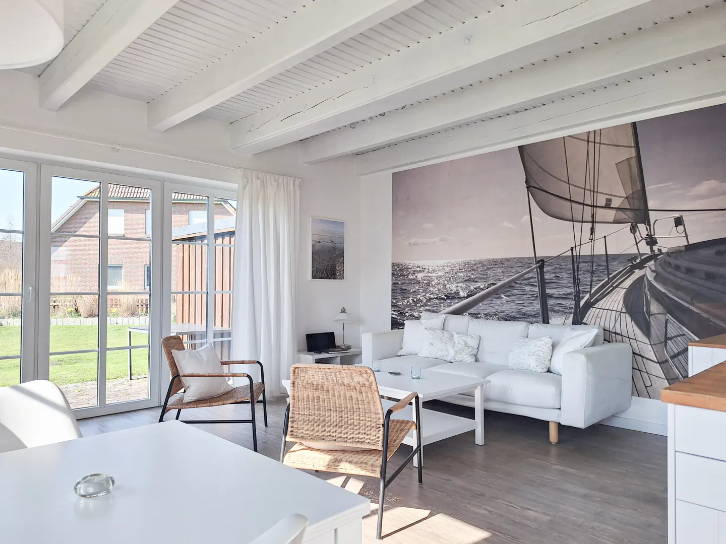 Bright living room with white walls, wood floors, and a white beamed ceiling. A large sailboat mural adorns one wall, and wicker chairs accent the space.