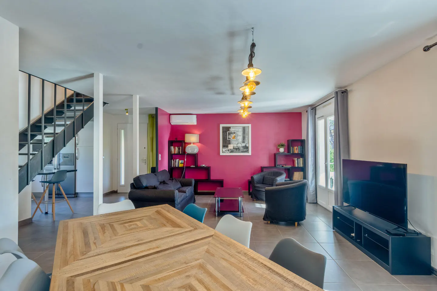 Open-concept living space with a wooden dining table, black sofa, and a bright pink accent wall with bookshelves. A staircase is visible.