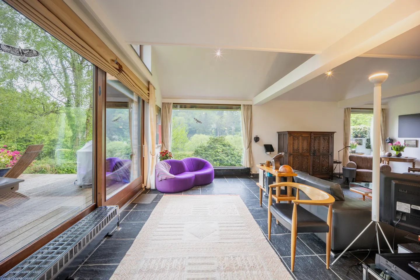 Bright living room with black tile floor, large windows overlooking green trees, and a purple sofa.