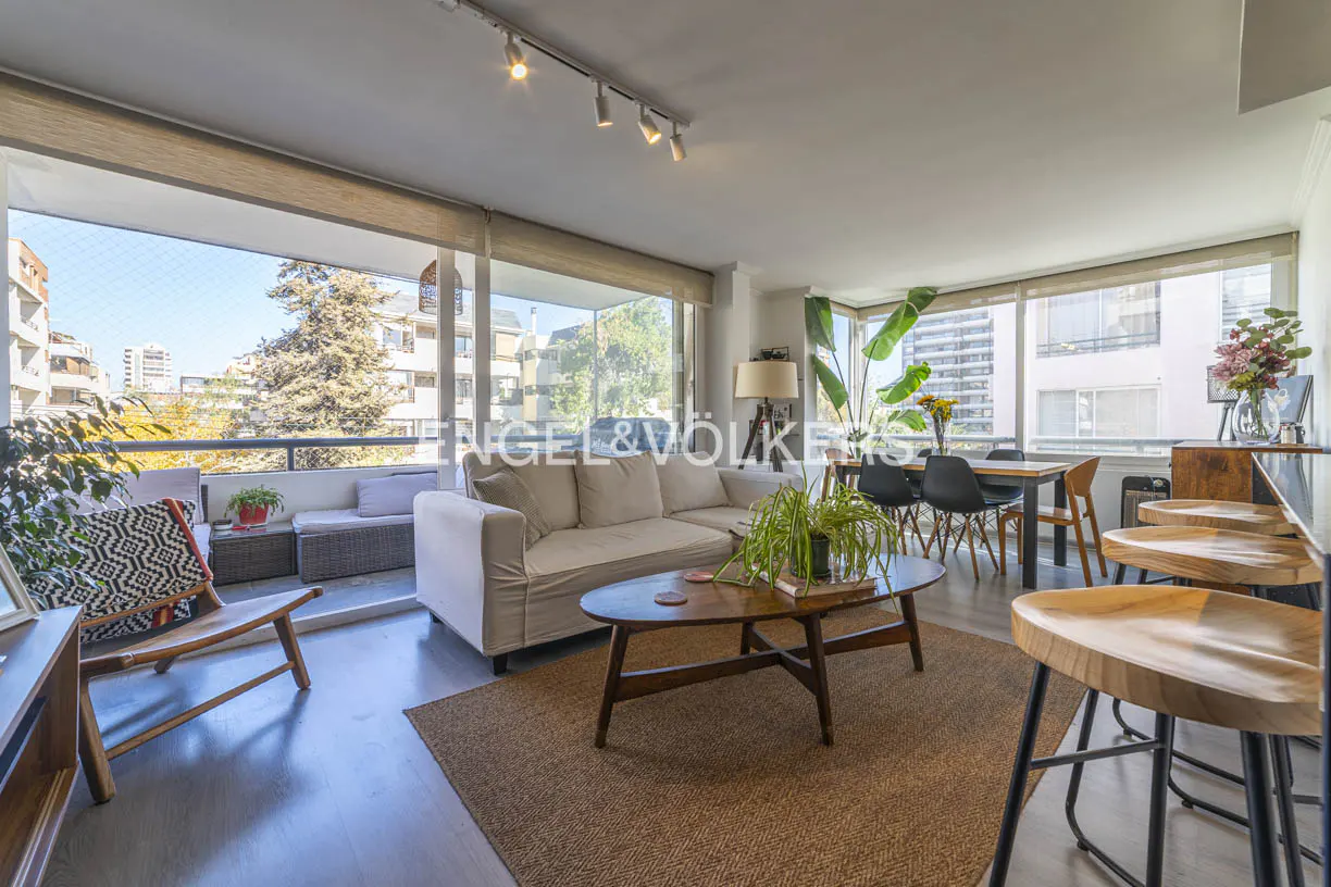 Bright living room with a beige sofa, wood coffee table, and stools. A balcony with seating overlooks a cityscape. Engel & Völkers logo visible.