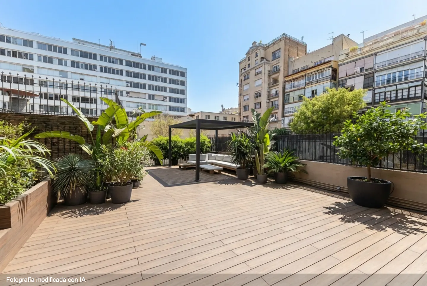 Wide shot of a rooftop terrace with wood flooring, plants, and a pergola with white seating. Buildings are visible in the background.