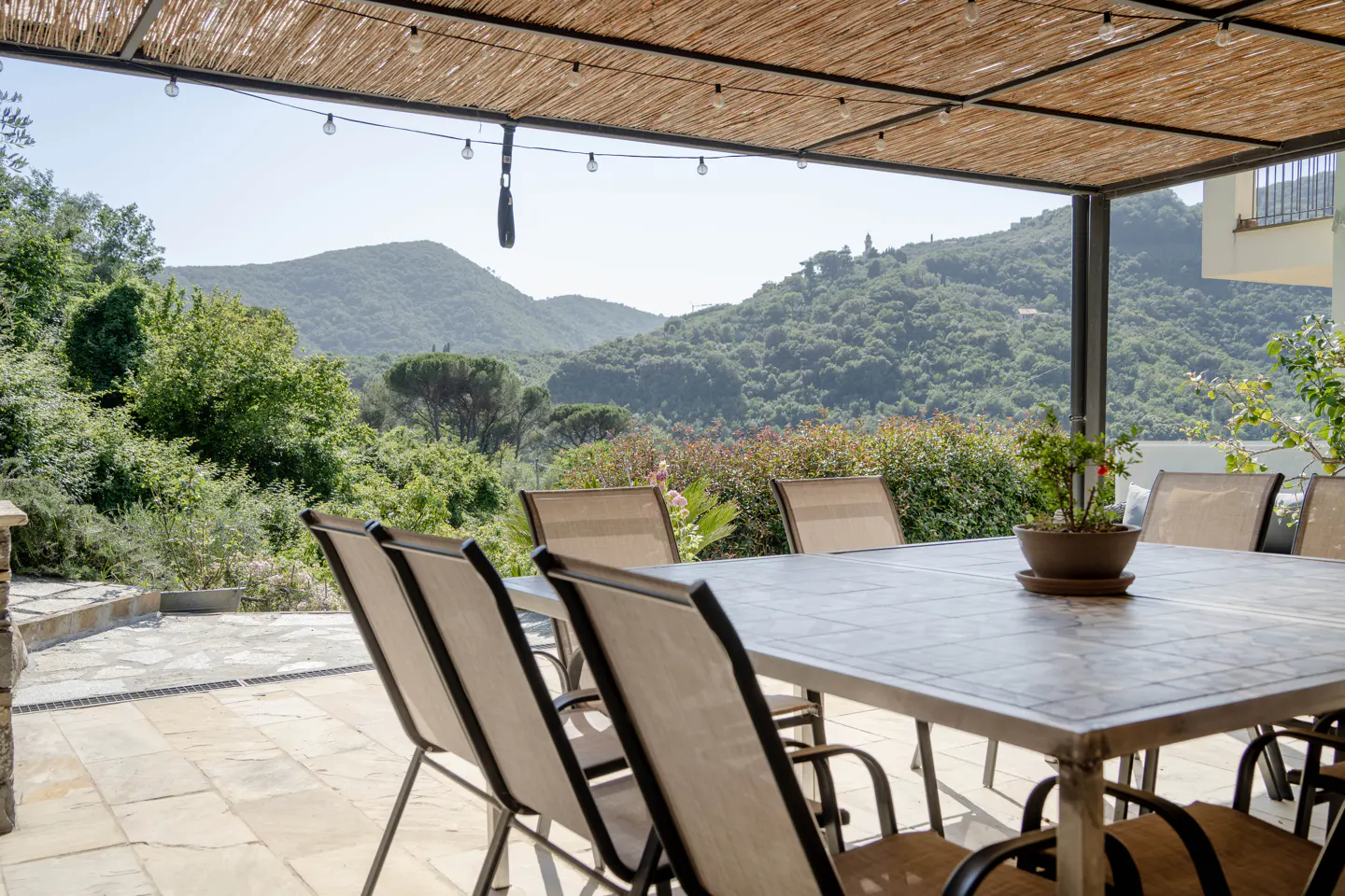 Outdoor patio with a table, chairs, and a view of green hills. A straw roof covers the patio, with string lights hanging.