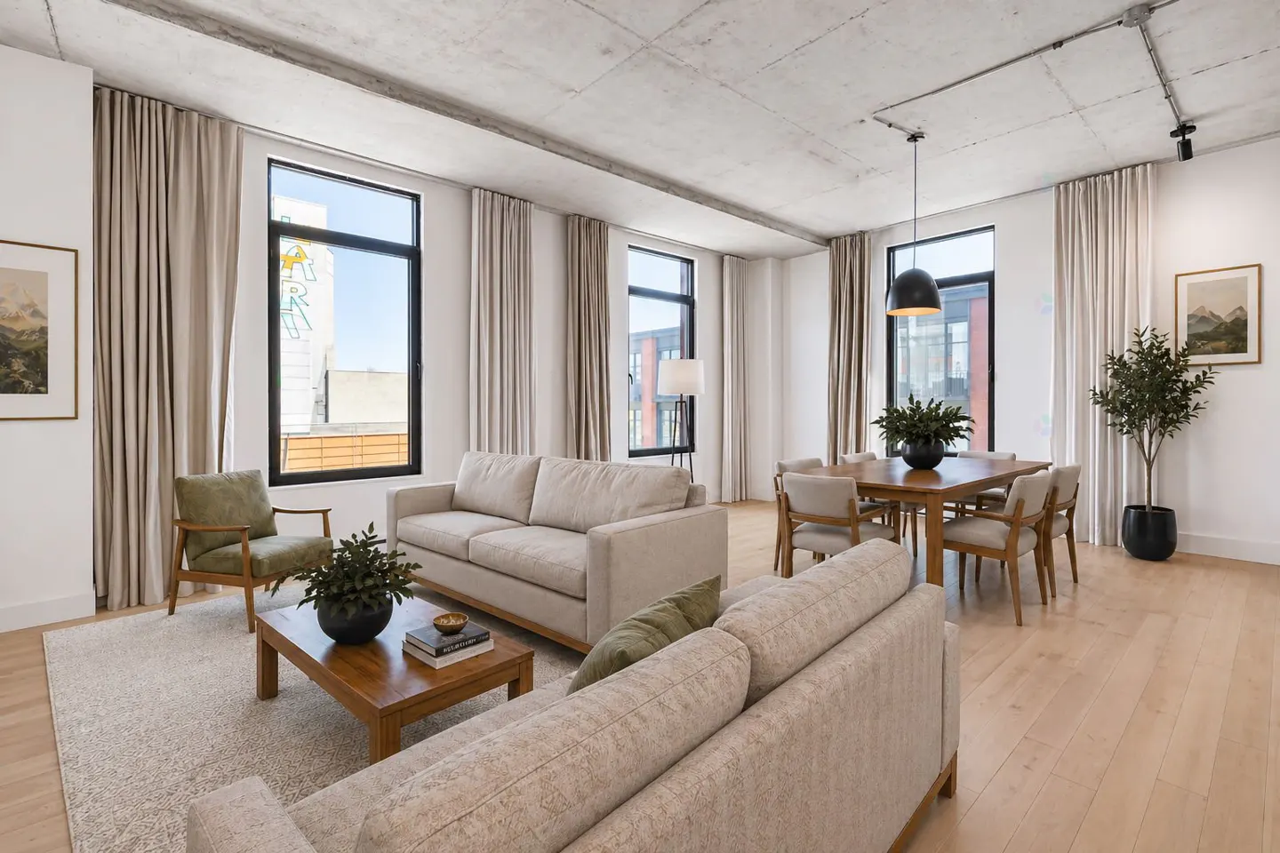 Bright, modern living room with two beige sofas, wood floors, and large windows with beige curtains. A dining table with chairs sits near a potted tree.