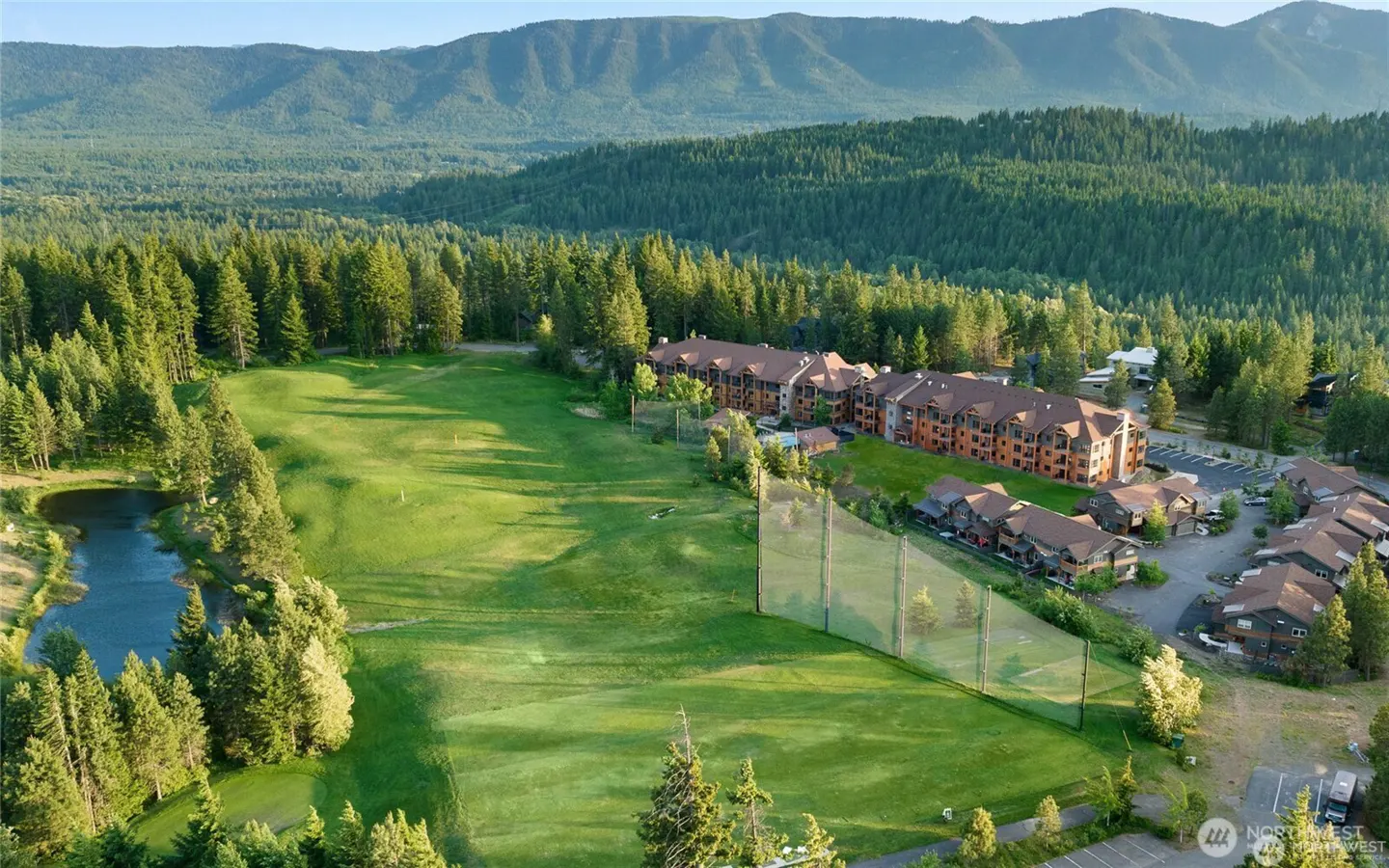 Aerial view of a golf course with a pond, driving range net, and multi-story resort buildings nestled in a pine forest.