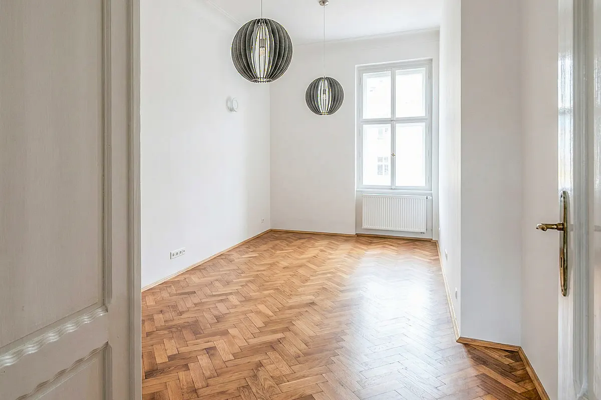 Bright, empty room with herringbone wood floors, white walls, and two globe pendant lights. A window with a radiator is visible.