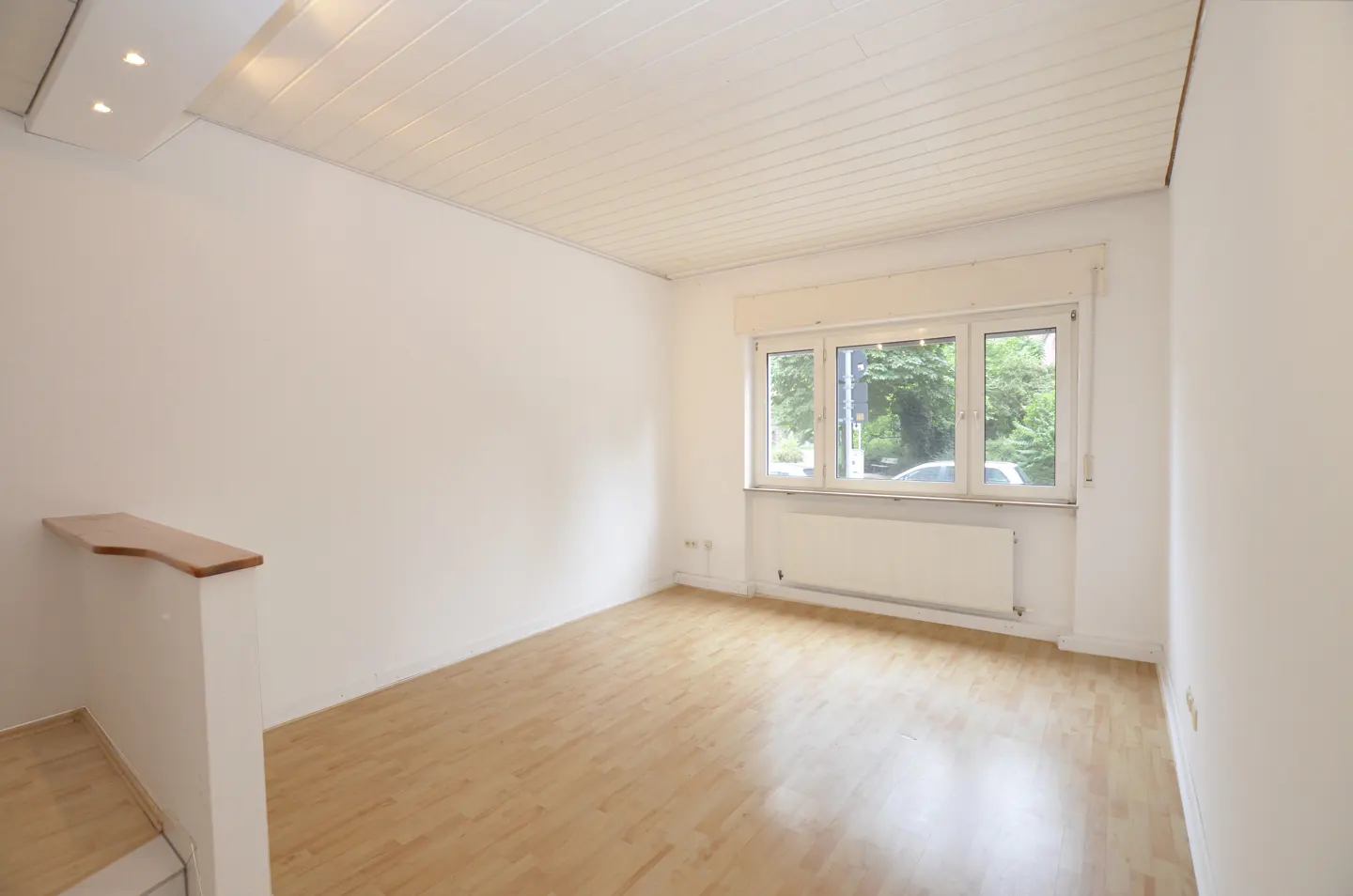 Bright, empty room with wood floors, white walls, and a large window showing trees and a car outside.