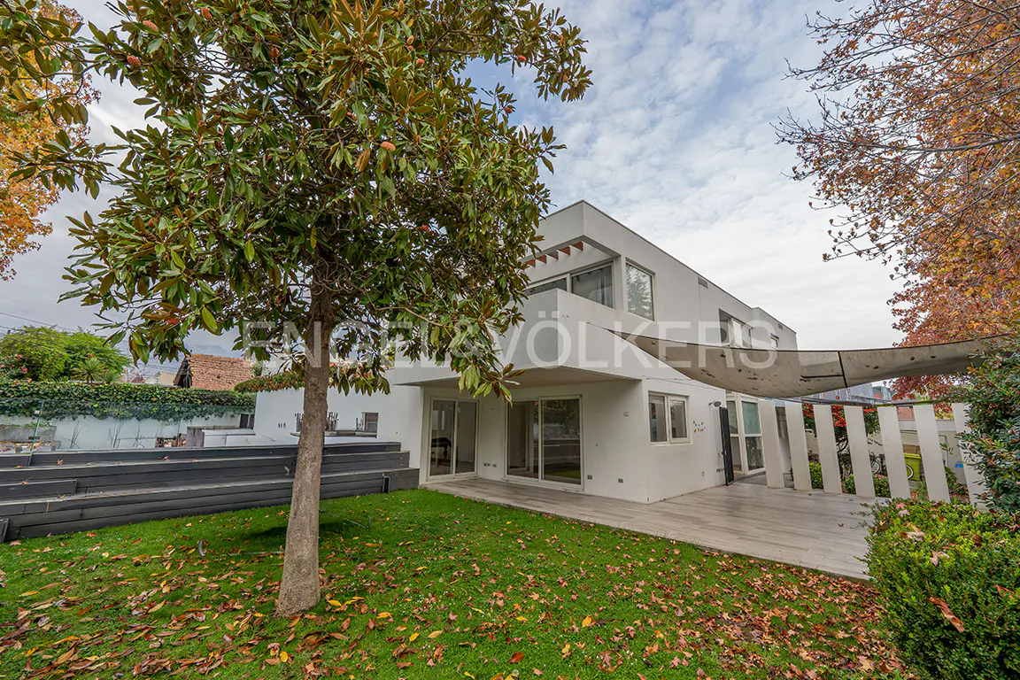 Exterior view of a modern white two-story house with a green lawn and trees. Fallen leaves are scattered on the grass.
