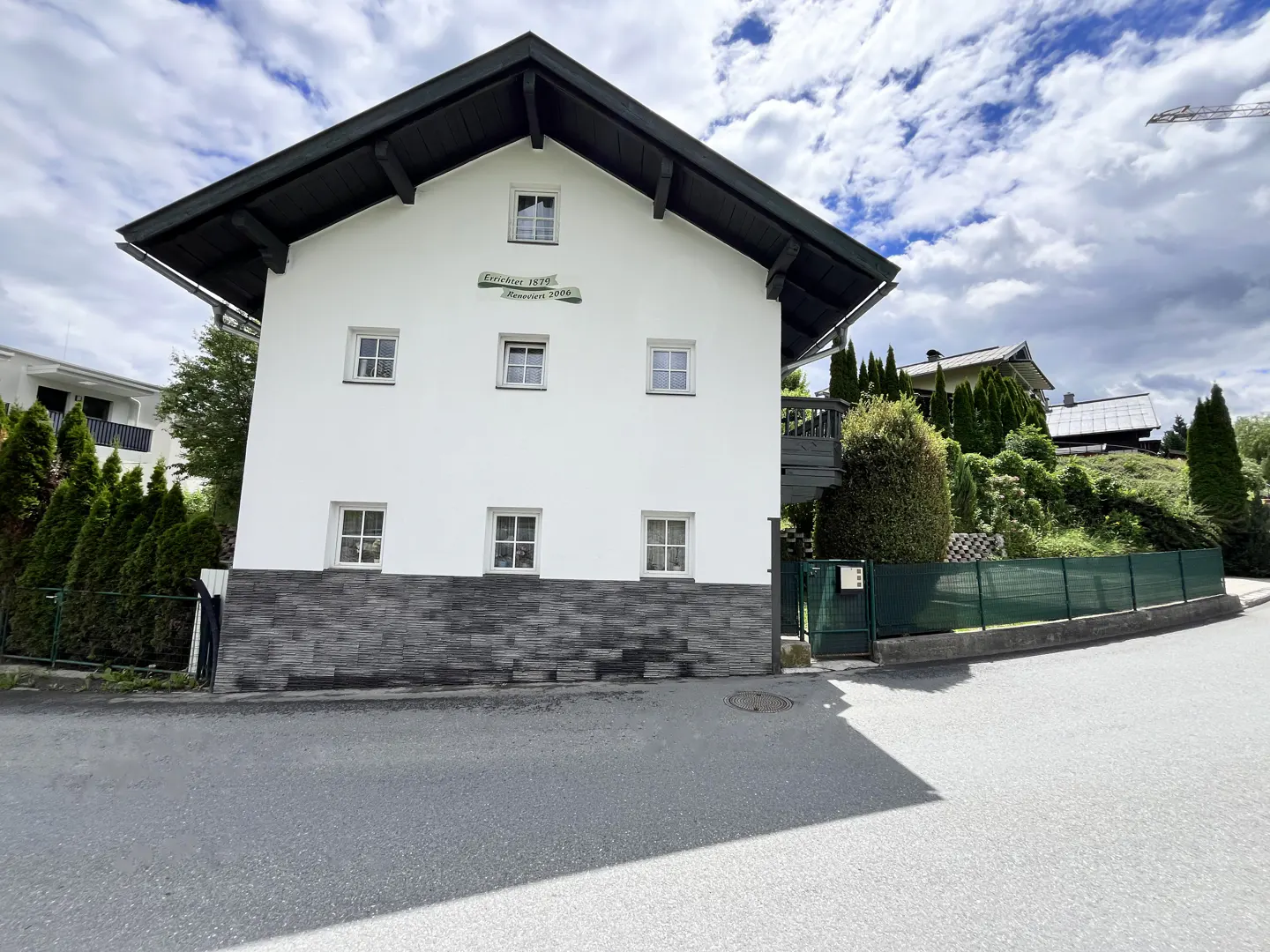 A white house with a dark roof and stone base, featuring small windows and a green fence, set against a cloudy sky.