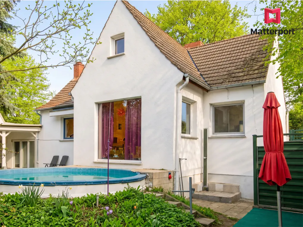 Exterior view of a white house with a brown roof, a round pool, and a red umbrella.