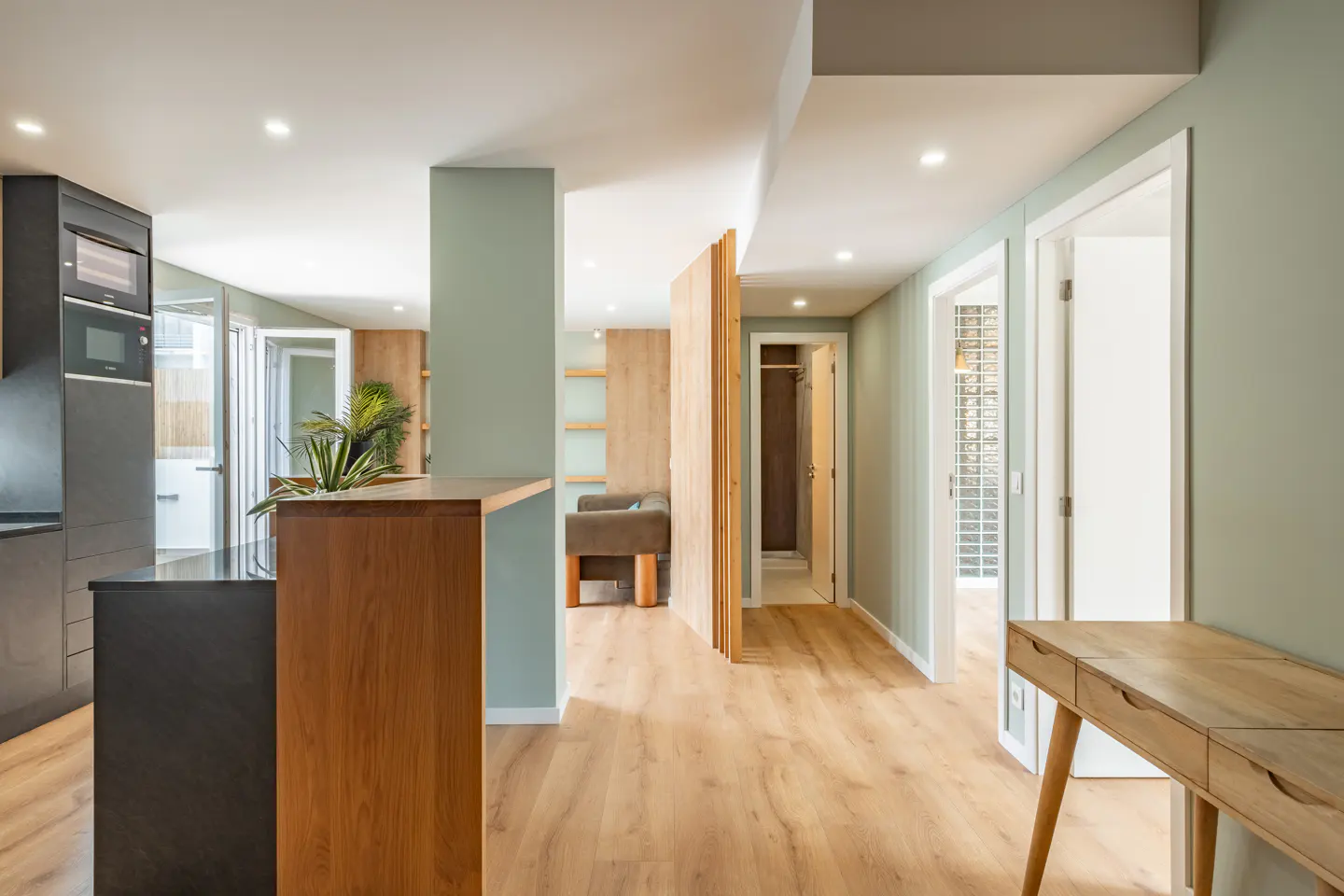 Interior view of a modern apartment with wood floors, sage green walls, and a black kitchen island. Hallway leads to other rooms.