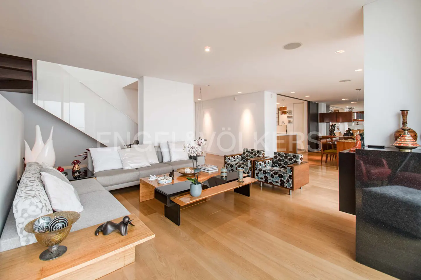 Bright, modern living room with wood floors, gray sofa, patterned chairs, and a wood coffee table. A staircase is visible on the left.
