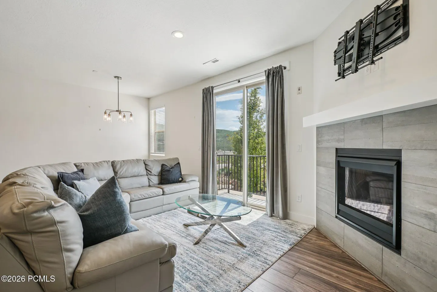 Living room with gray leather sectional, glass table, fireplace, and balcony with mountain view.