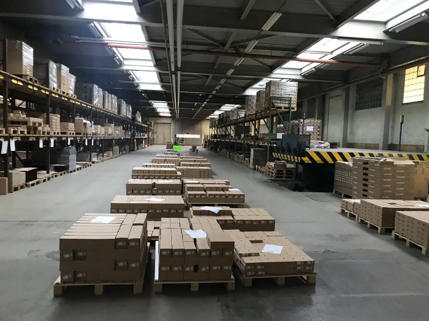 A warehouse interior with rows of cardboard boxes on pallets, metal shelving, and a loading dock with yellow and black stripes.