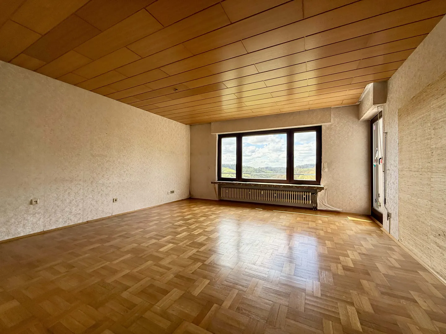 Empty room with parquet floor, wood paneled ceiling, textured walls, and a large window with a view.