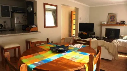 A bright living room with a round wooden table covered in a colorful plaid cloth, chairs, and a view of the kitchen and living area.