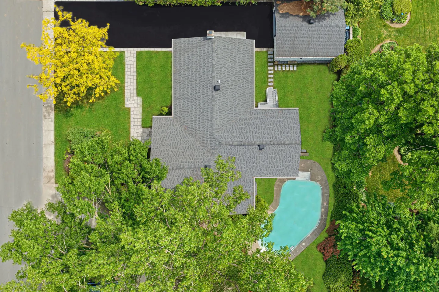 Aerial view of a house with a gray roof, a backyard pool, lush green trees, and a paved driveway.