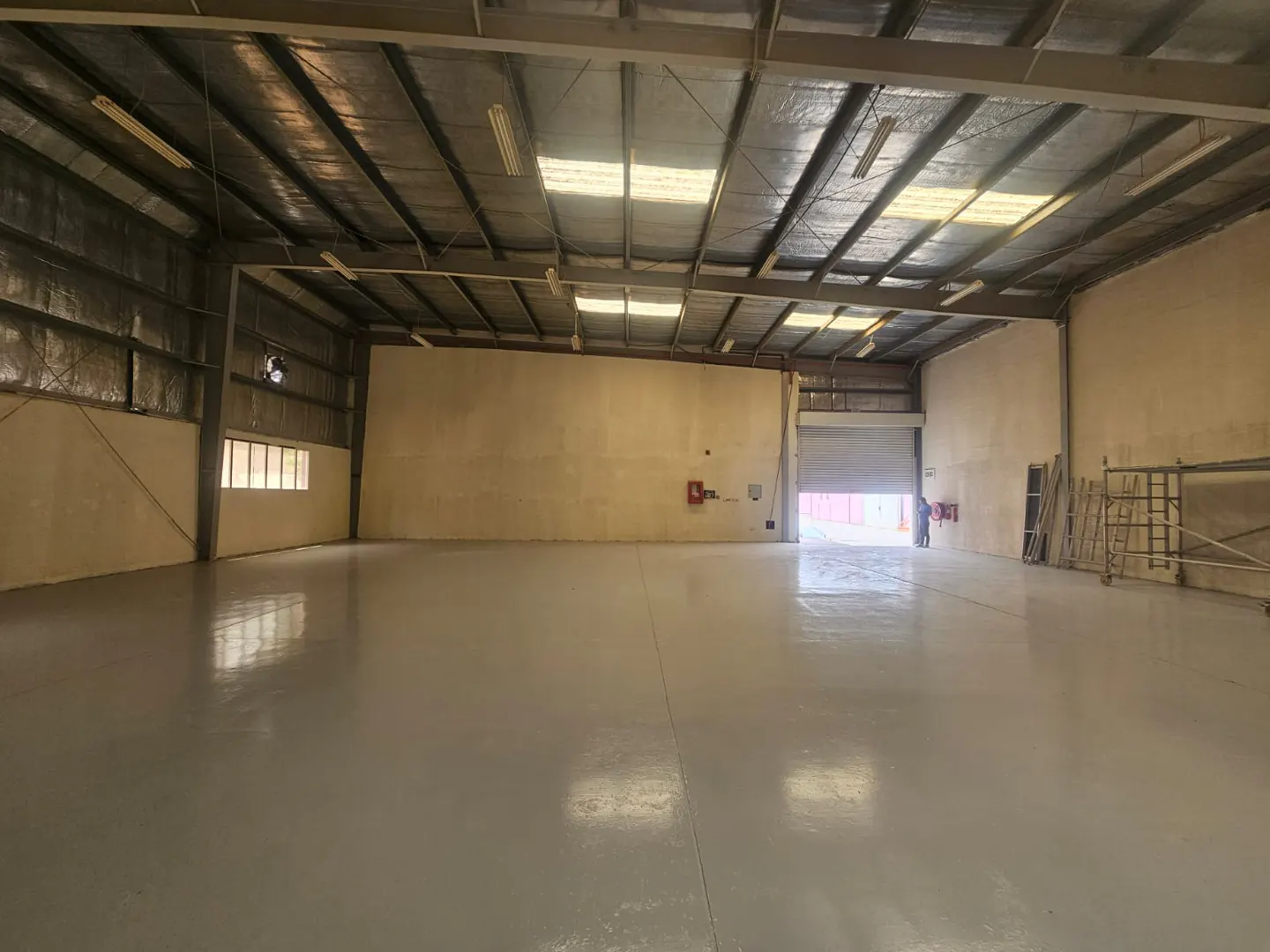 A large, empty warehouse with a gray floor, beige walls, and a high, metal ceiling with fluorescent lights.