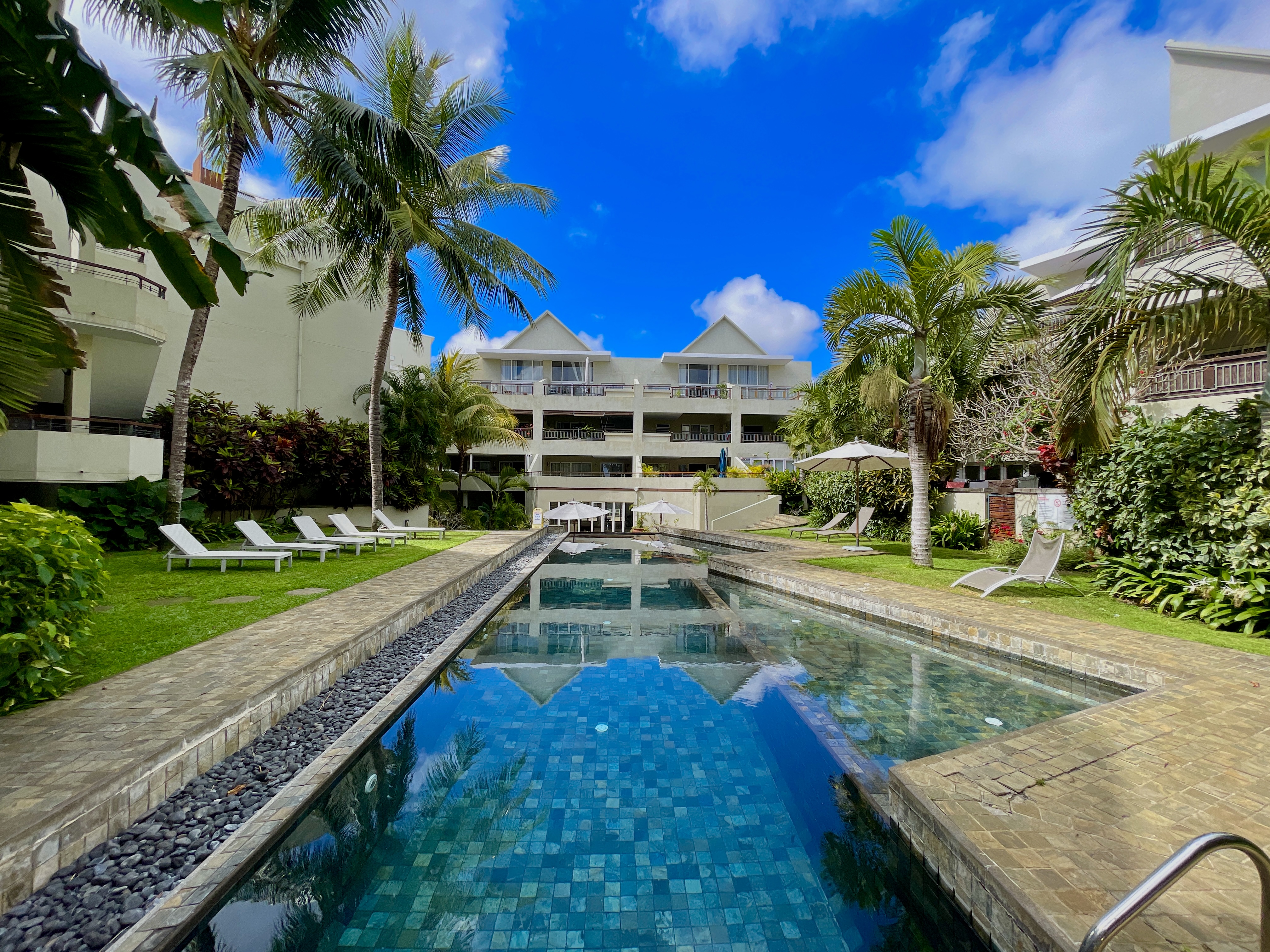 Long rectangular swimming pool surrounded by palm trees, green lawns, lounge chairs, and a multi-story resort building. [AI-generated text]