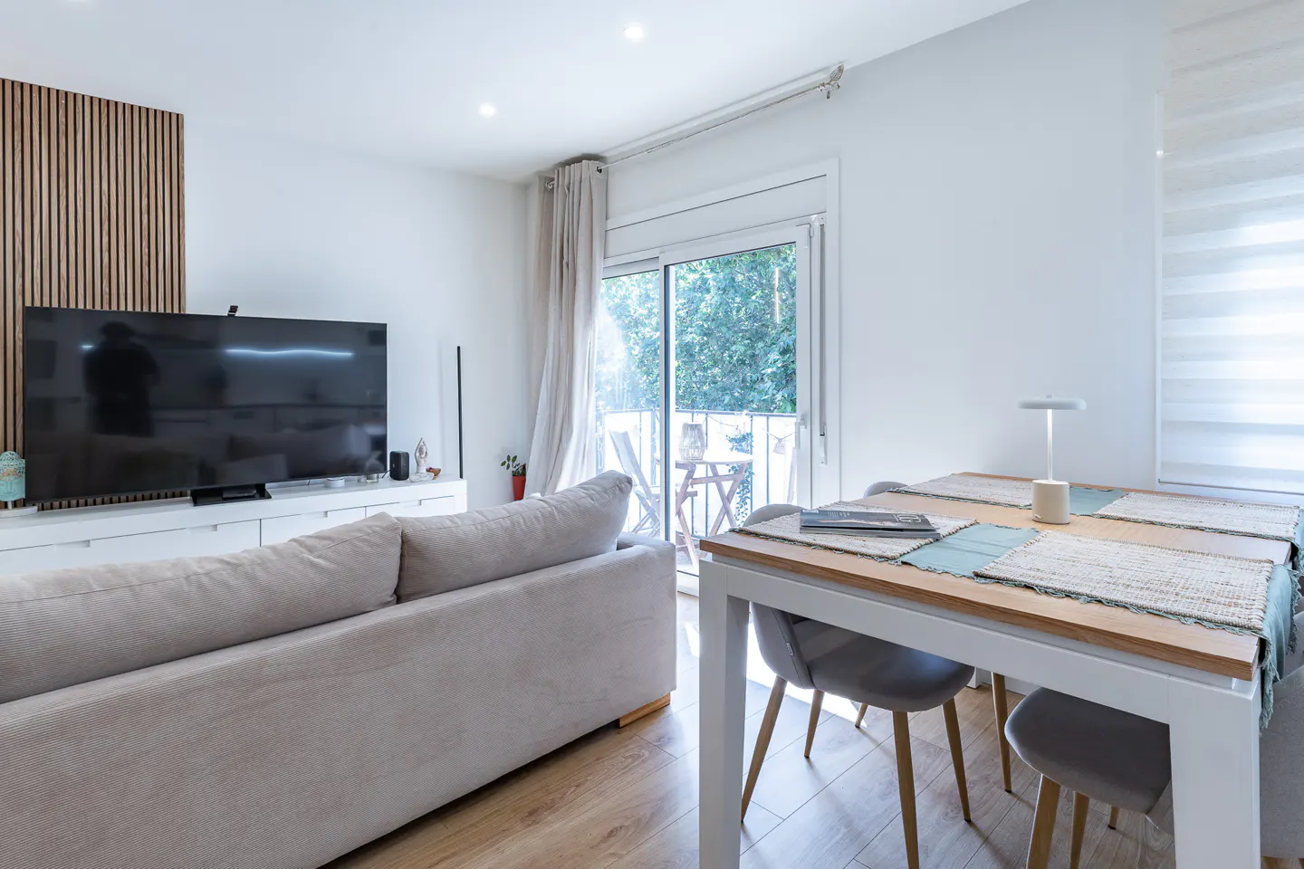 Bright living room with a beige sofa, a large TV, and a white dining table with gray chairs. Balcony with greenery visible through sliding glass doors.