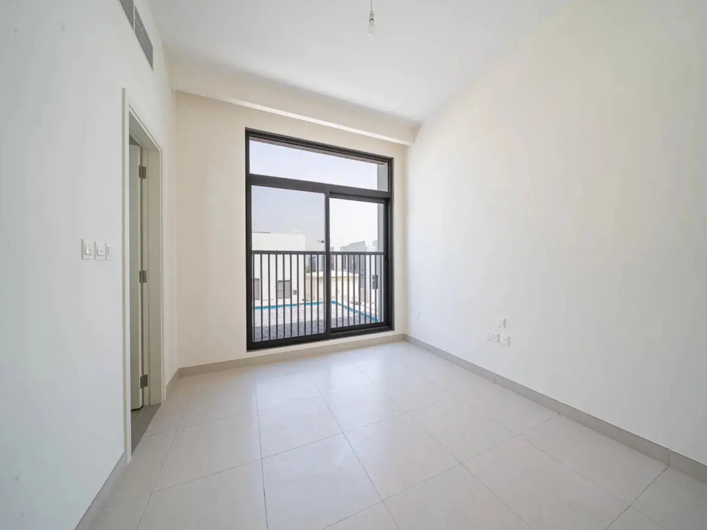 Bright, empty room with white walls and tile floor. A black-framed sliding glass door leads to a balcony overlooking a pool.