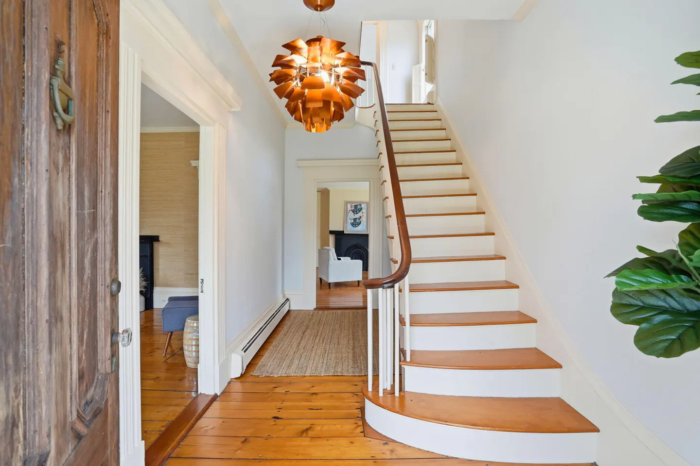 Open wood door reveals a foyer with wood floors, white walls, and a staircase. A modern copper light fixture hangs above.
