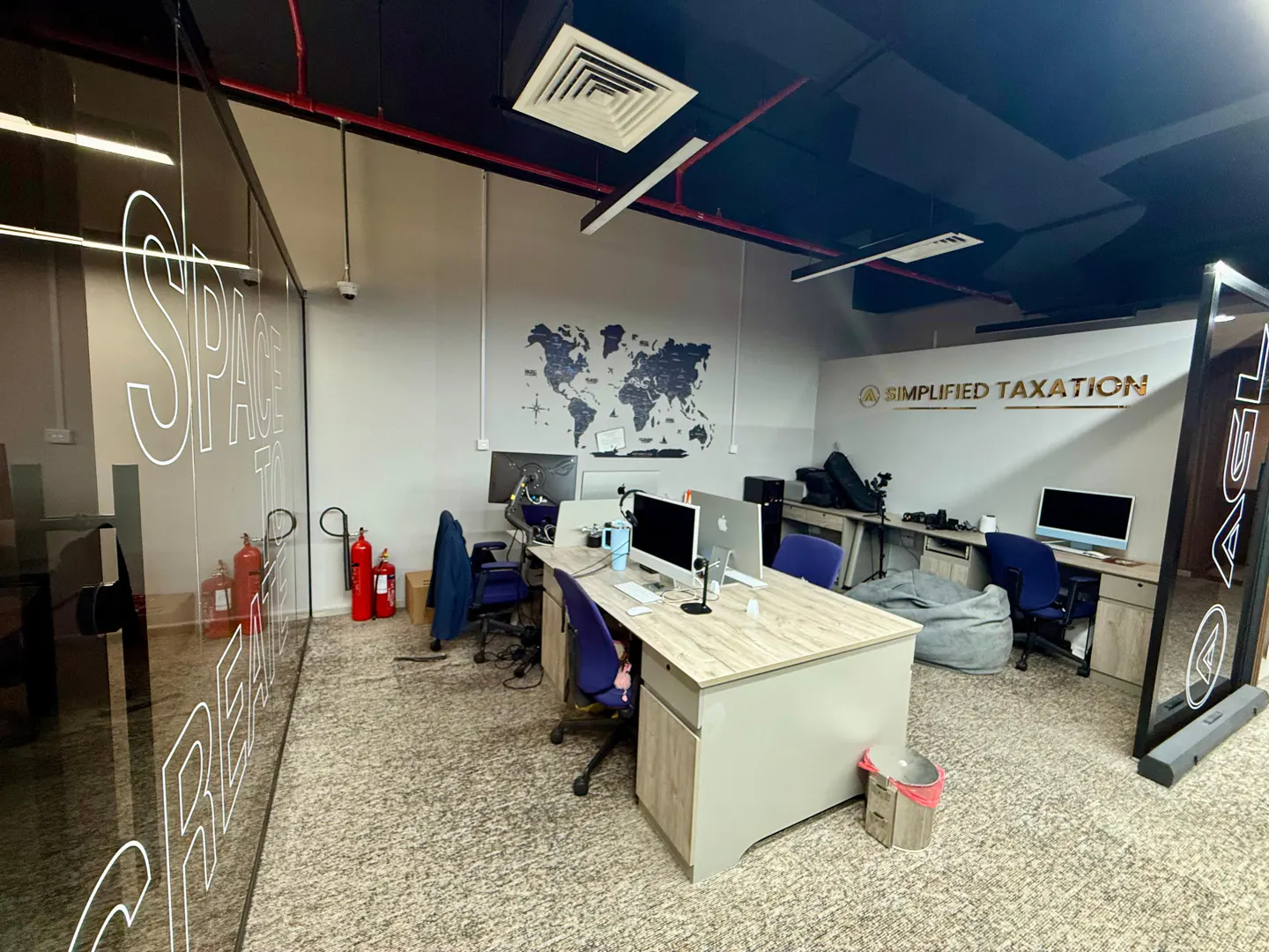 Office space with desks, computers, and chairs. A world map hangs on the wall. "Simplified Taxation" is displayed above a desk. Two red fire extinguishers are visible.
