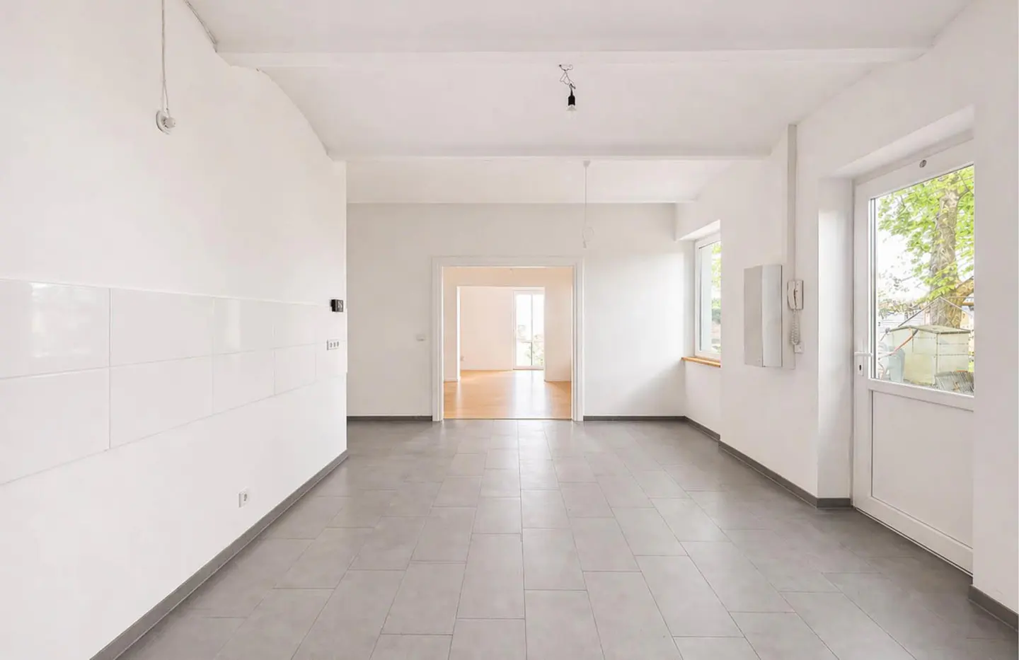 Bright, empty room with white walls, gray tile floor, and natural light from windows and a door to the outside.