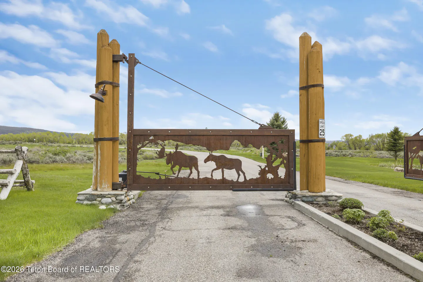 Rustic metal gate with moose silhouettes, supported by wooden posts on stone bases, against a green field and blue sky.