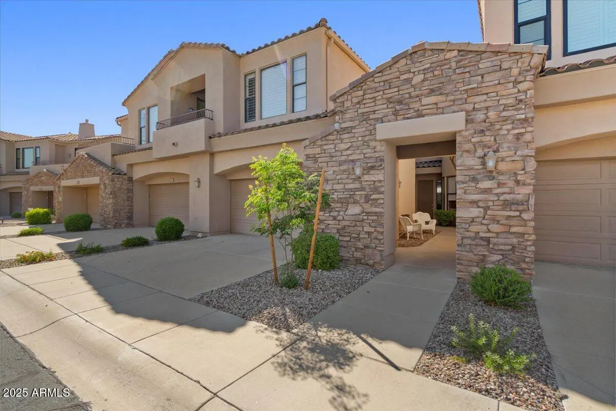 Exterior of a beige two-story townhouse with stone accents, a balcony, and a two-car garage. A small tree is planted in front.