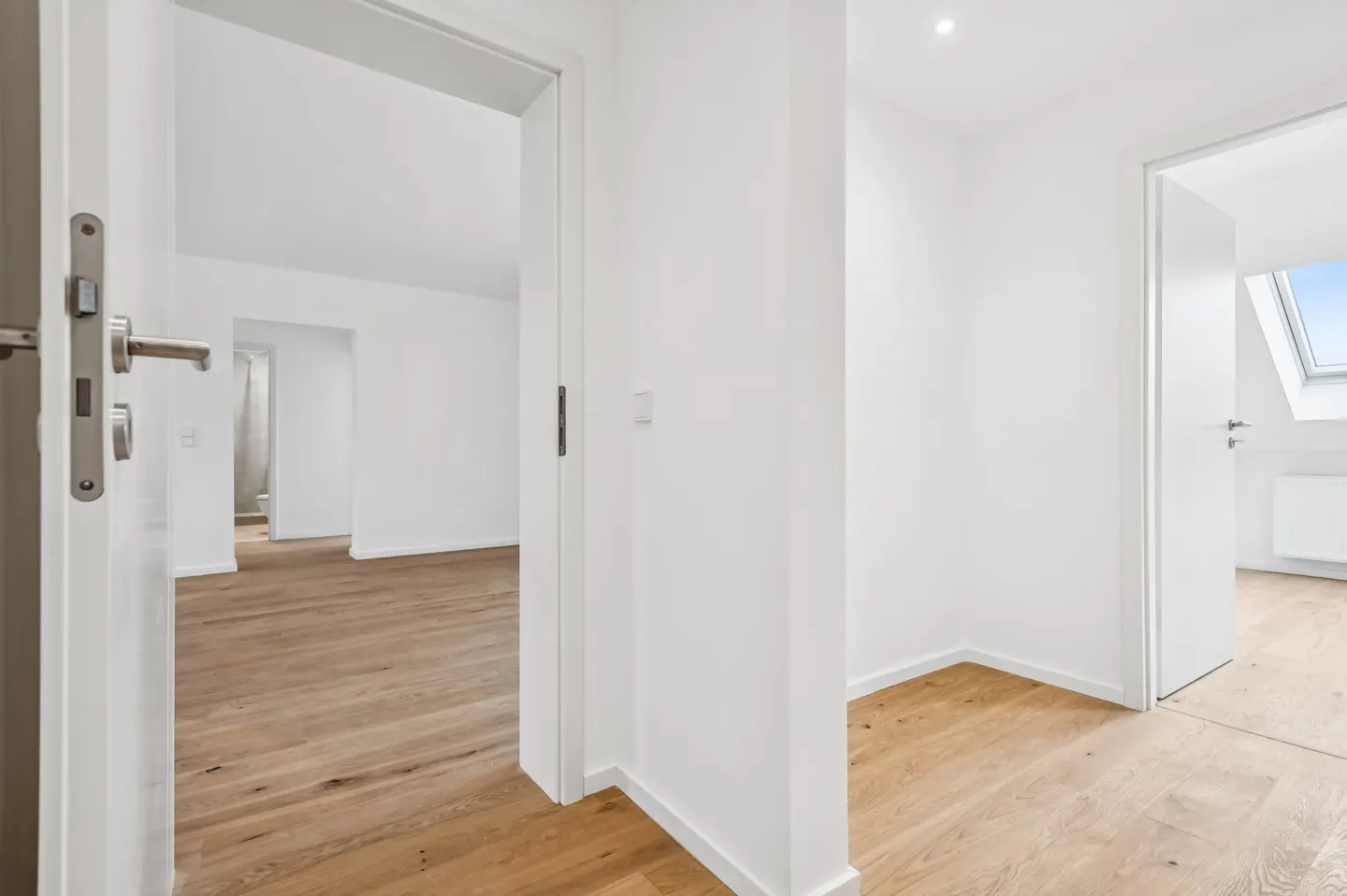 Bright hallway with white walls, light wood floors, and open doorways leading to multiple rooms, including one with a skylight.