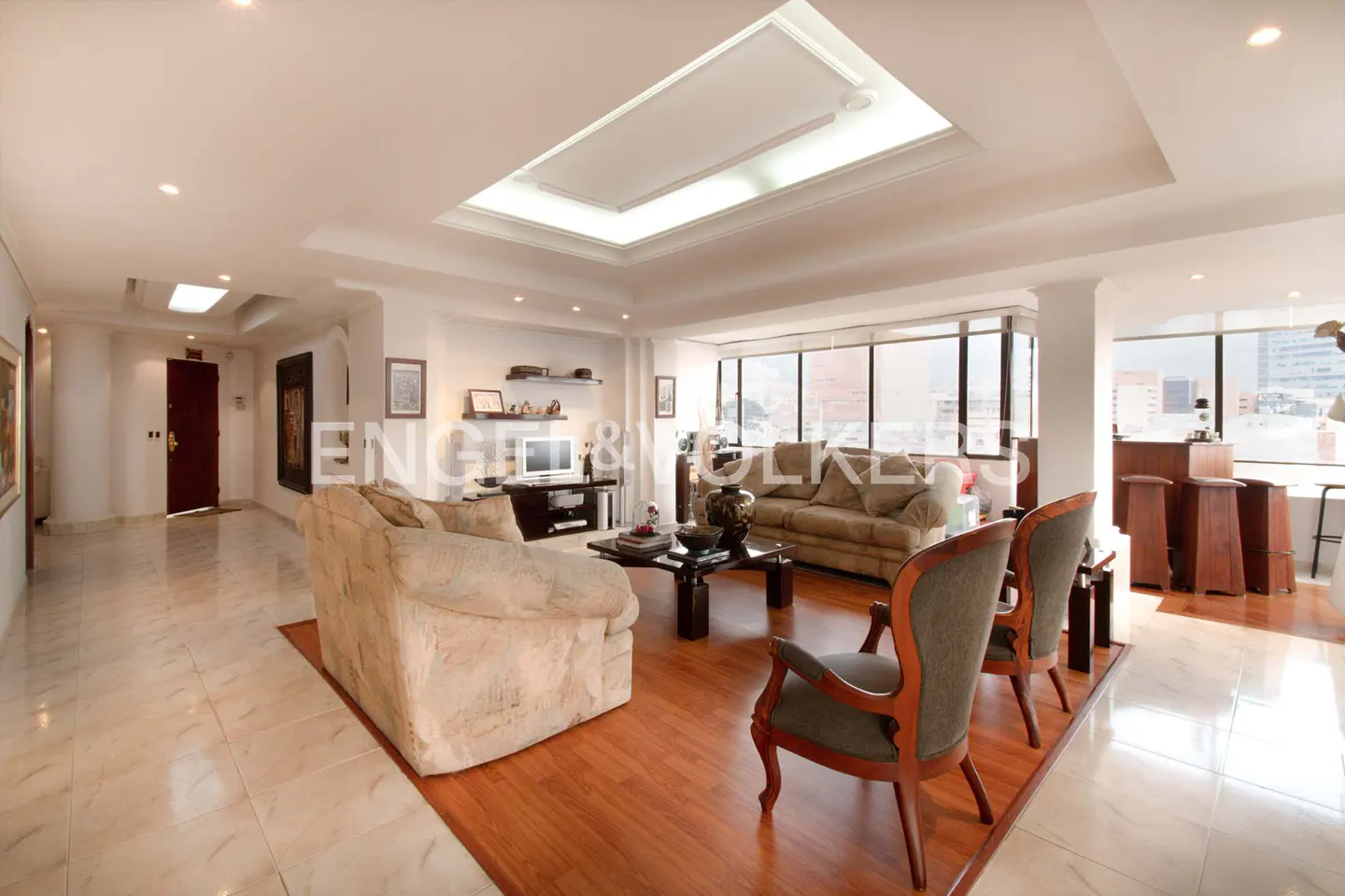 Bright living room with beige sofas, wooden chairs, and a black coffee table on a wood floor. Large windows offer city views.