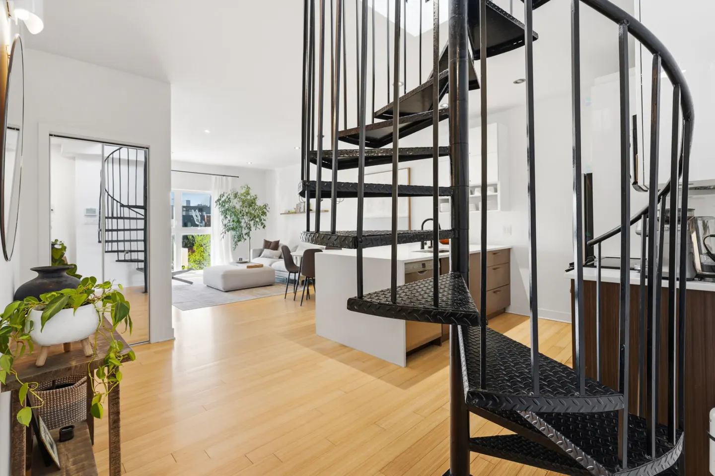 Bright, modern interior with a black spiral staircase. Living room and kitchen visible in the background. Wooden floors and white walls.