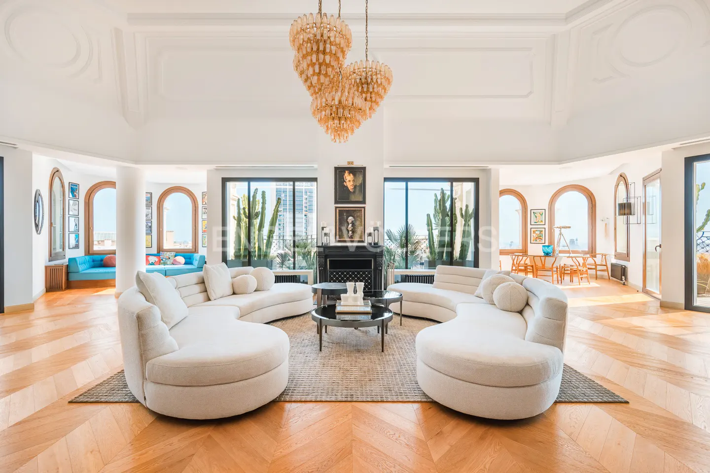 Bright, spacious living room with curved white sofas, herringbone wood floors, and a large chandelier. Windows offer city views.