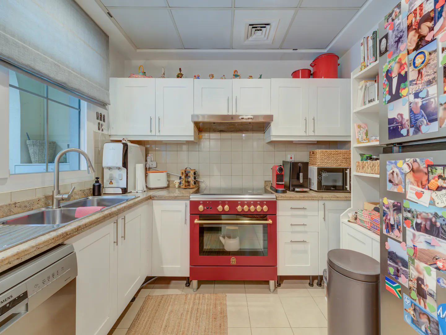 Bright kitchen with white cabinets, granite countertops, and a red oven. A window provides natural light. Refrigerator covered in photos.