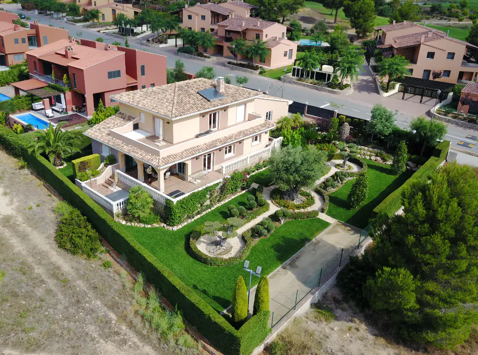 Aerial view of a two-story beige house with a tile roof, surrounded by a green hedge, manicured lawn, and other houses.