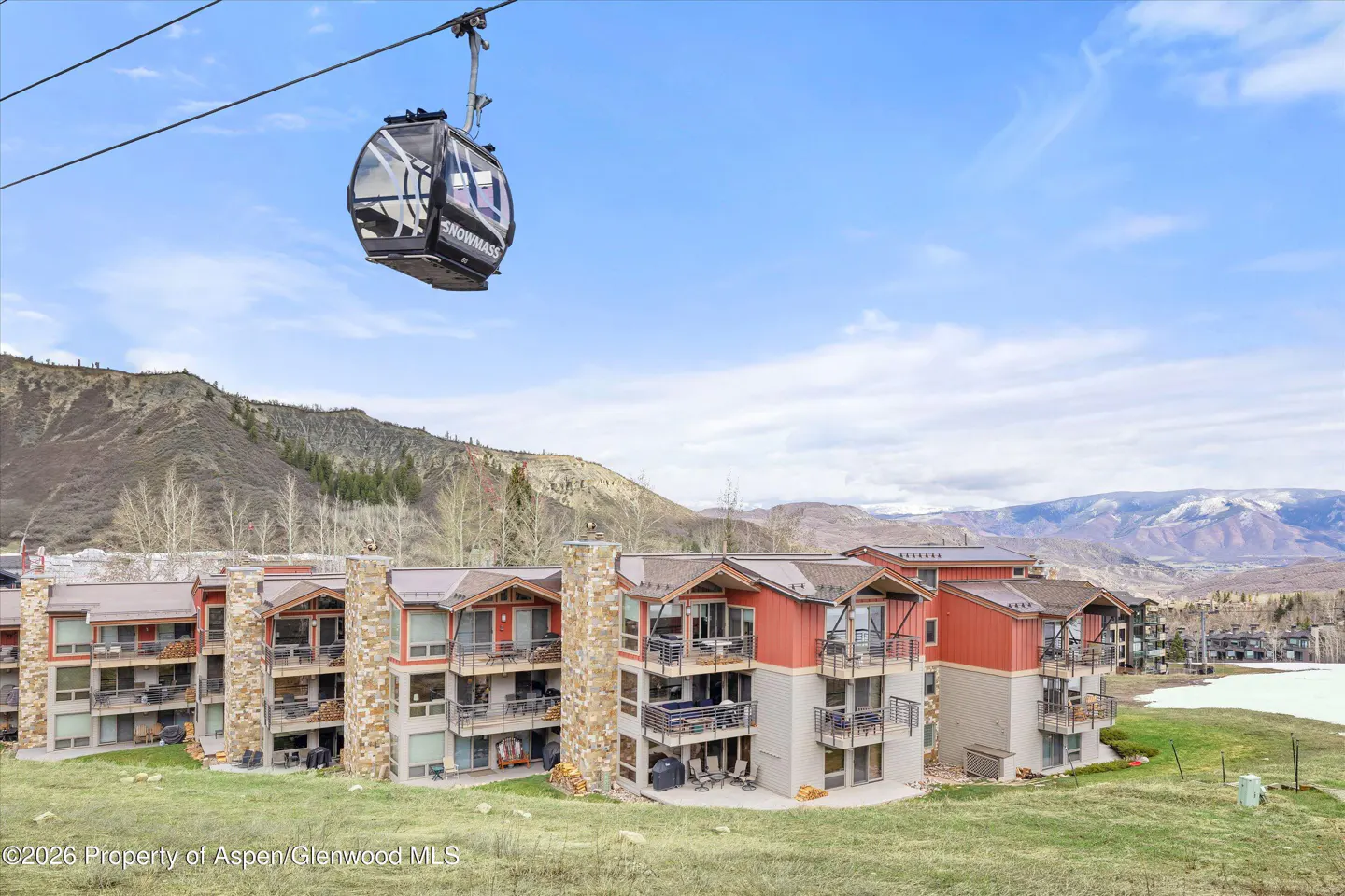 Condos with balconies sit below a Snowmass gondola against a mountain backdrop. The buildings are red and gray with stone accents.