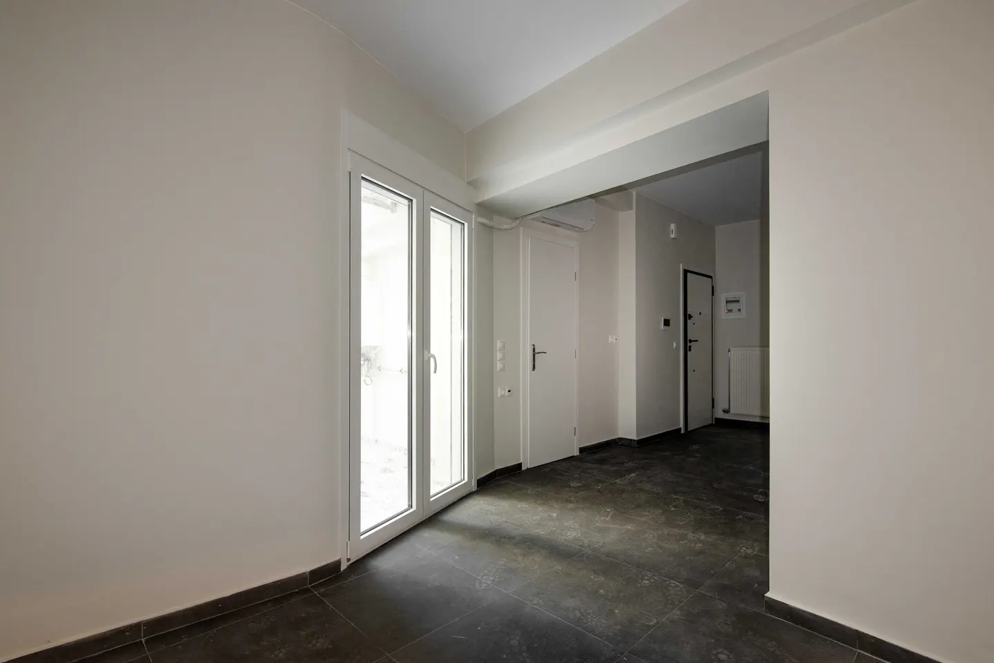 A bright, empty room with white walls, dark tile floor, and a white-framed glass door to the outside.