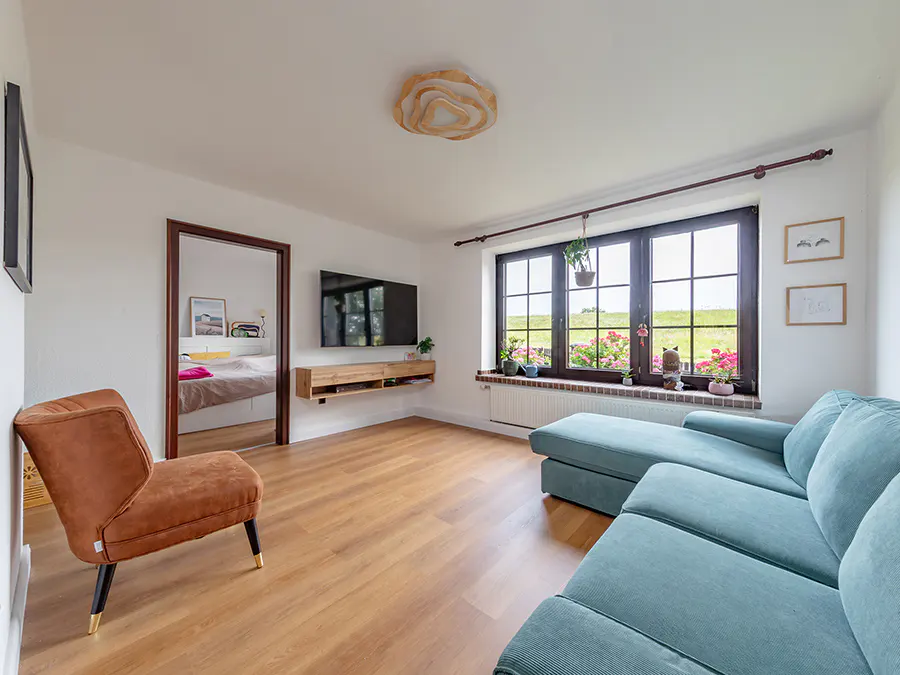 Bright living room with a blue sofa, brown chair, and a large window overlooking a green field. A TV is mounted above a wooden shelf.